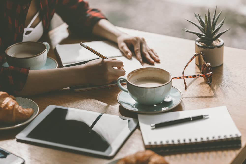 A hand with a pencil over a notebook, a tablet and a pale blue cup of coffee